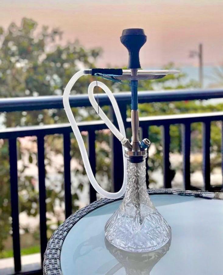 A clear Shisha set on a round glass-top table with a woven base, featuring a white hose, a silver metal stem, and a black mouthpiece, photographed outdoors with a balcony railing and blurred background.